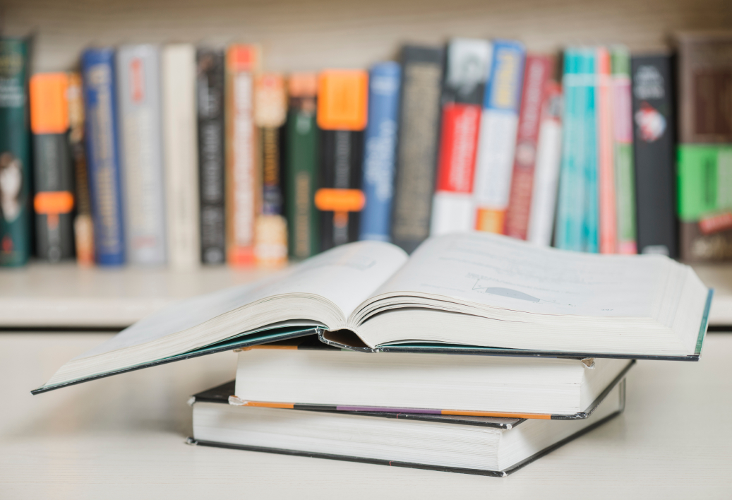 stack of books rests on a table, with a bookshelf filled with more books in the background
