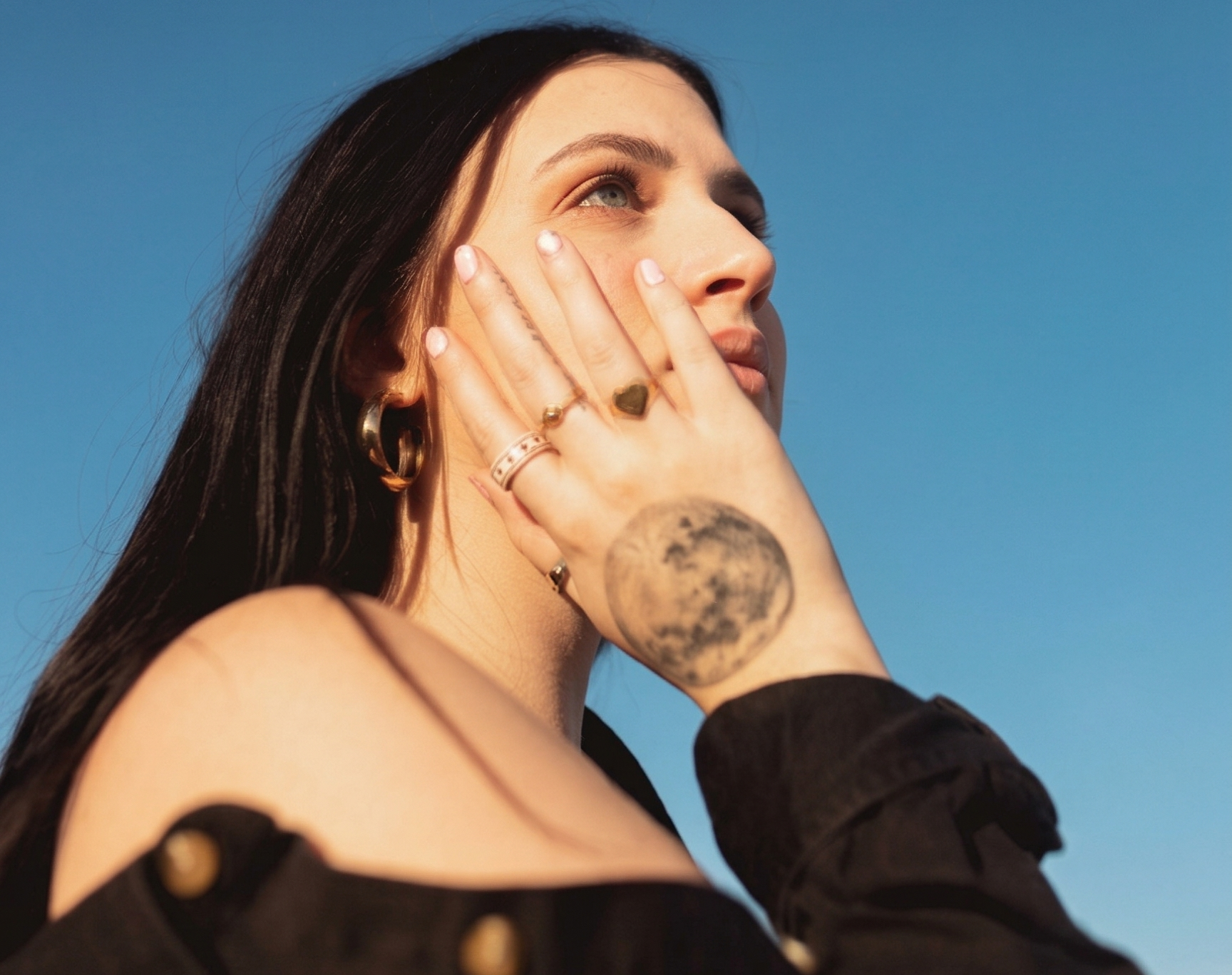 Woman with moon tattoo on hand, gazing at sky, wearing black off-shoulder top and hoop earrings, clear blue background